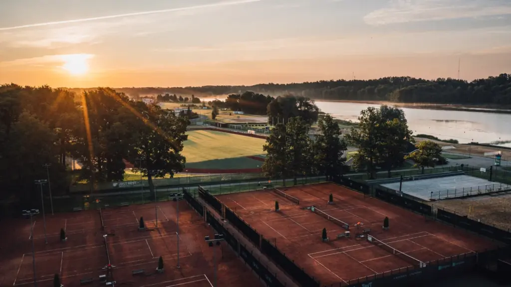 Vaade ülevalt tenniseväljakutele päikeseloojangus, taustal on staadion, puud ja rahulik järv.